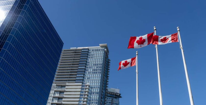 Three Canadian flags next to buildings against a blue sky.